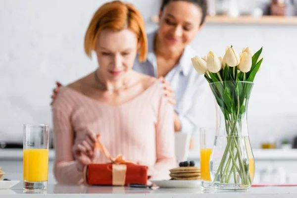 Africaine américaine lesbienne femme étreignant épaules de petite amie ouvrir boîte cadeau près du petit déjeuner et vase avec tulipes, flou au premier plan — Photo de stock