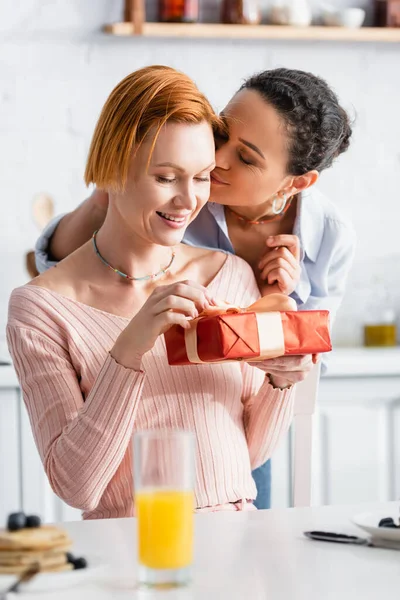 Africaine américaine lesbienne femme baisers copine ouverture cadeau boîte sur valentines jour dans cuisine, flou au premier plan — Photo de stock