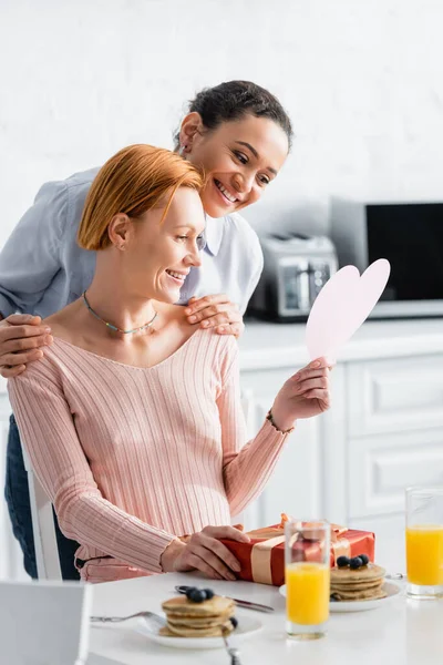Smiling african american woman hugging shoulders of happy lesbian girlfriend holding paper cut heart on valentines day — Stock Photo