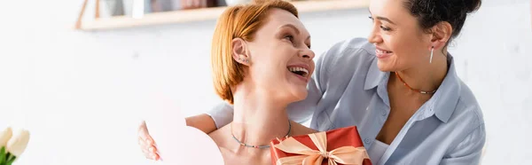 Amazed redhead woman with gift box looking at african american lesbian girlfriend, banner — Stock Photo