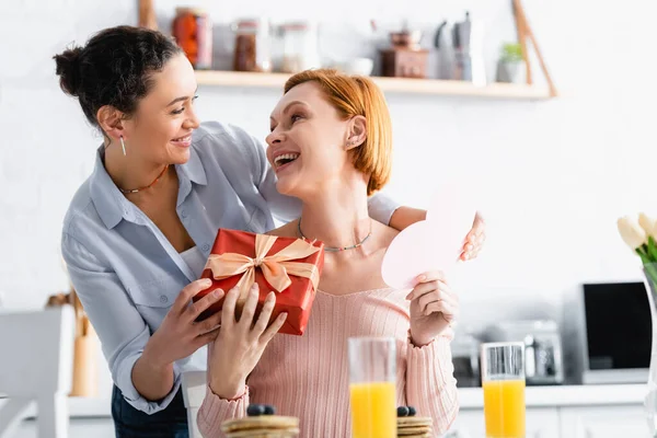 Happy african woman presenting gift box and paper cut heart to astonished lesbian girlfriend, blurred foreground — Stock Photo