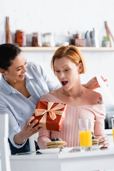 Cheerful african american woman holding paper cut heart and gift box near amazed lesbian girlfriend, blurred foreground — Stock Photo