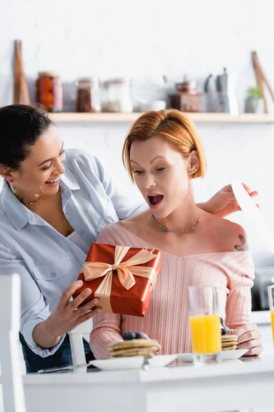 Fröhliche afrikanisch-amerikanische Frau mit Herz aus Papier und Geschenkschachtel in der Nähe der erstaunten lesbischen Freundin, verschwommener Vordergrund — Stockfoto