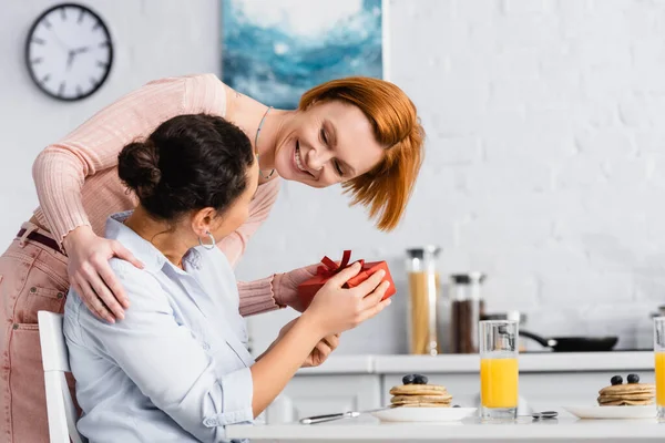 Alegre lesbiana mujer presentando regalo en valentines día a africano americano lesbiana novia en cocina - foto de stock