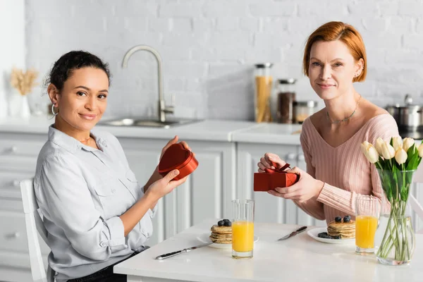 Glückliches interrassisches lesbisches Paar blickt in die Kamera, während es Geschenke beim Frühstück am Valentinstag hält — Stockfoto
