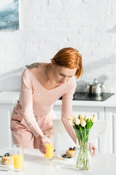 Redhead tattooed woman serving breakfast on table near tulips in kitchen — Stock Photo