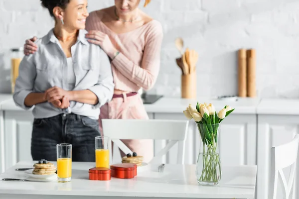 Focus selettivo della tavola con colazione, regali di San Valentino e tulipani vicino coppia lesbica interrazziale su sfondo sfocato — Foto stock