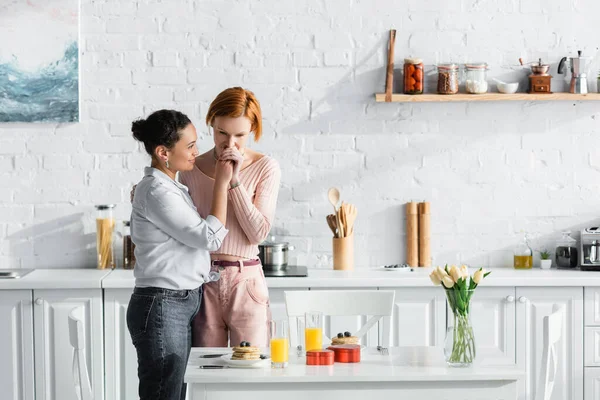 Lesbienne femme baisers main de afro-américaine petite amie près de la table avec Saint Valentin cadeaux et petit déjeuner dans la cuisine moderne — Photo de stock