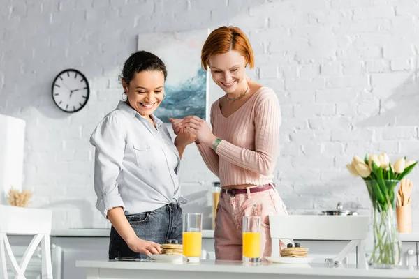 Happy interracial lesbian couple holding hands near table with breakfast and tulips — Stock Photo