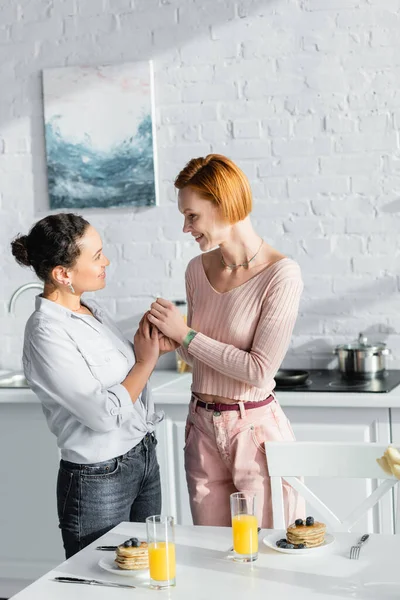 Smiling interracial lesbian couple holding hands and looking at each other near breakfast on table — Stock Photo