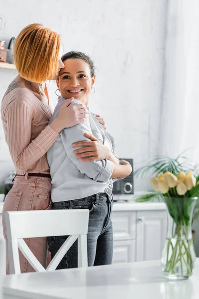 Redhead lesbian woman embracing shoulders of happy african american girlfriend in kitchen — Stock Photo