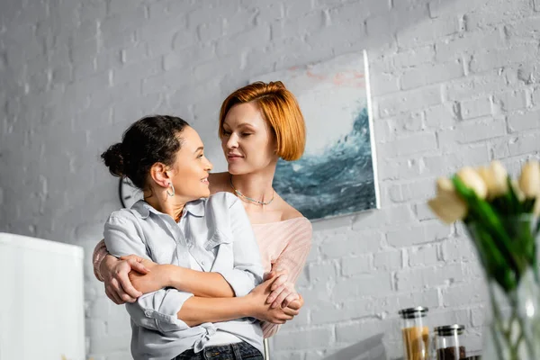 Happy interracial lesbian couple embracing and looking at each other in kitchen on blurred foreground — Stock Photo