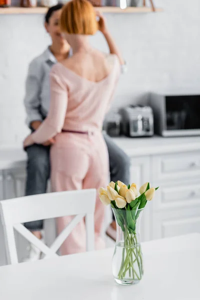 Selective focus of tulips in vase on kitchen table near interracial lesbian couple on blurred background — Stock Photo