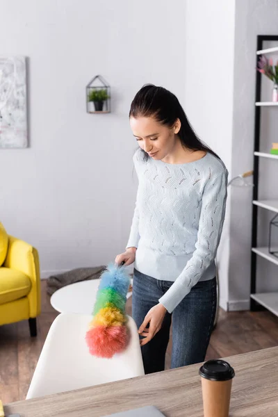 Joven esposa limpieza tabla de planchar con cepillo de polvo cerca de café para ir a la mesa - foto de stock