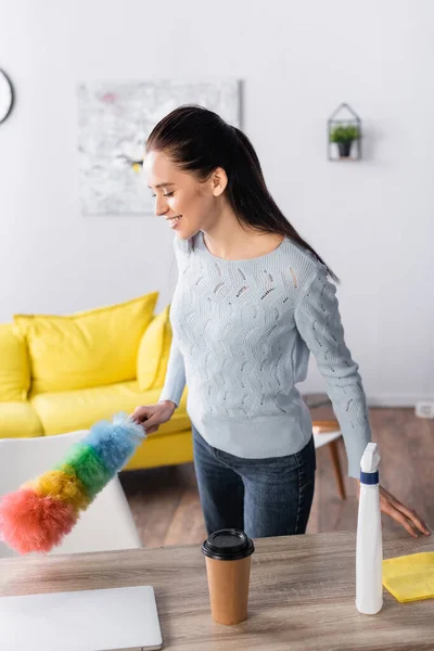 Young woman holding dust brush near detergent spray and coffee to go on table — Stock Photo