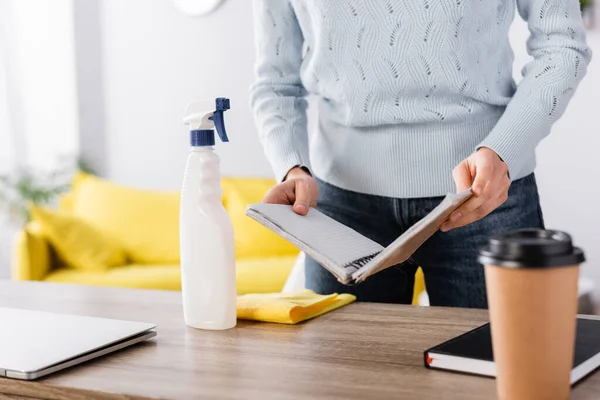 Partial view of housewife holding notebook near rag, detergent, and coffee to go on blurred foreground — Stock Photo