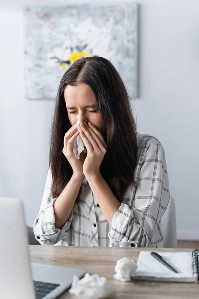 Freelancer estornuda en servilleta de papel mientras sufre de alergia cerca de la computadora portátil en primer plano borroso - foto de stock