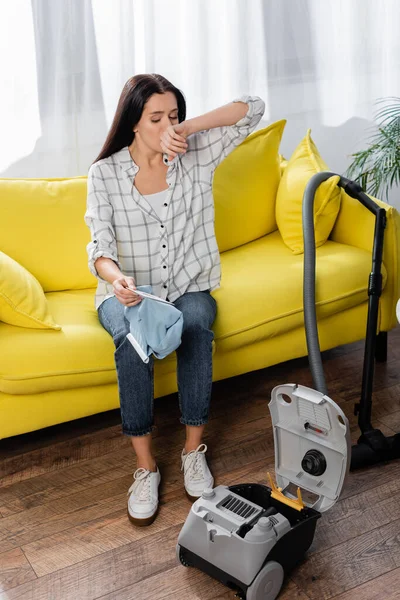 Allergic woman wiping nose while sitting on sofa with vacuum cleaner dust bag — Stock Photo