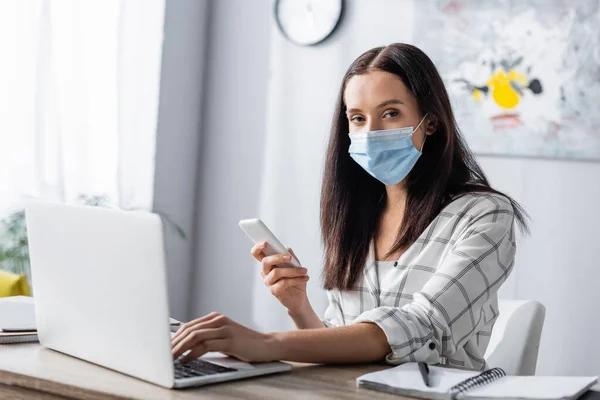 Freelancer in medical mask holding smartphone, typing on laptop and looking at camera — Stock Photo