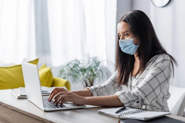 Freelancer en mascarilla médica escribiendo en laptop cerca de notebooks en casa - foto de stock
