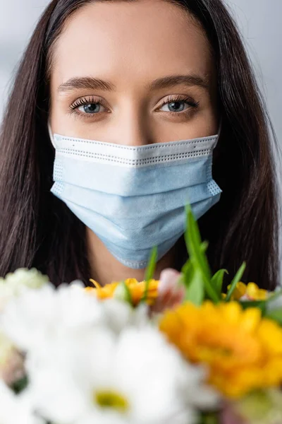 Portrait of allergic woman in medical mask looking at camera near flowers, blurred foreground — Stock Photo