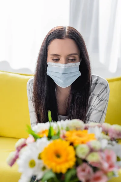 Allergikerin in medizinischer Maske sitzt neben Blumen auf verschwommenem Vordergrund — Stockfoto