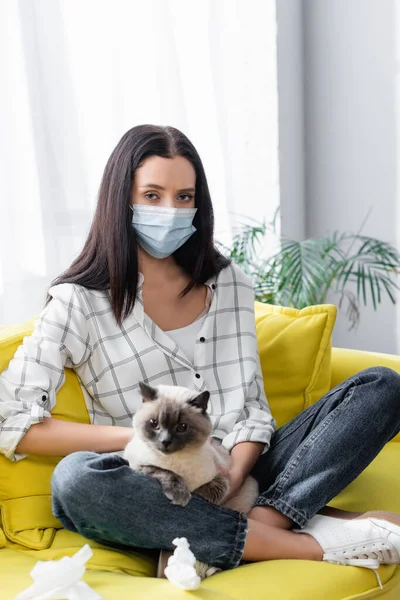 Алергічна жінка в медичній масці дивиться на камеру, сидячи на дивані з кішкою — стокове фото