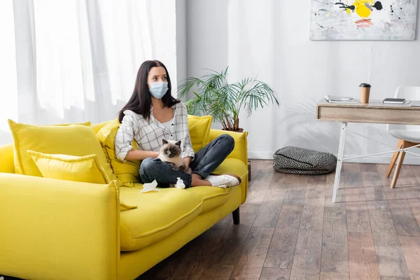 Femme allergique dans le masque médical assis avec chat sur canapé jaune — Photo de stock