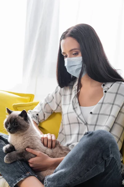Аллергик в медицинской маске обнимает кошку, сидя дома со скрещенными ногами — стоковое фото