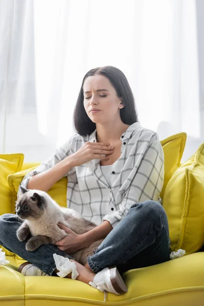 Mujer alérgica con los ojos cerrados tocando la garganta mientras está sentada en el sofá con el gato - foto de stock