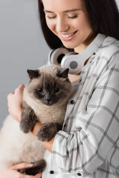 Mujer feliz con auriculares inalámbricos en el cuello sosteniendo gato en casa - foto de stock