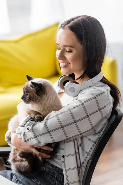 Mulher sorridente com fones de ouvido no pescoço abraçando gato em casa — Fotografia de Stock