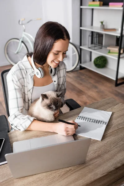 Happy freelancer writing in notebook while sitting near laptop with cat — Stock Photo