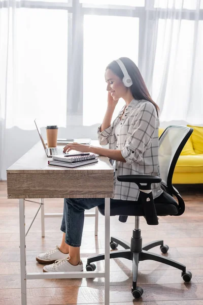 Side view of happy freelancer in headphones typing on laptop at home — Stock Photo