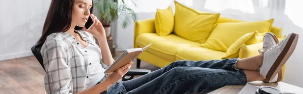 Freelancer sentado con las piernas en la mesa, sosteniendo el cuaderno y hablando por teléfono móvil, pancarta - foto de stock