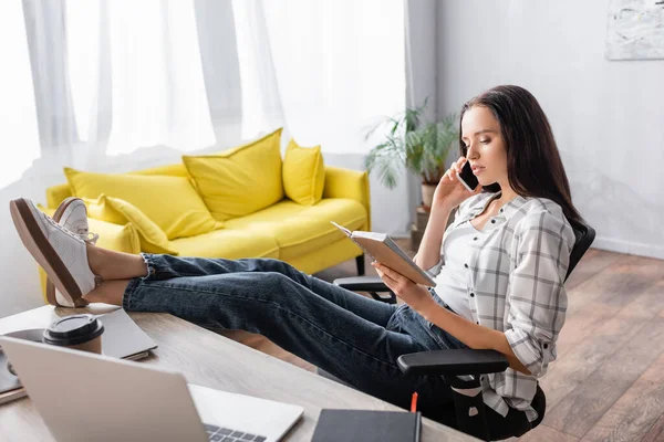 Freelancer sitting with legs on table, looking in notebook and talking on smartphone — Stock Photo