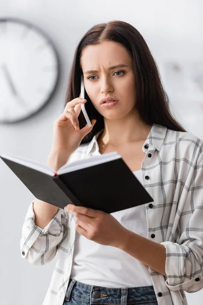 Worried freelancer holding notebook while talking on mobile phone at home — Stock Photo
