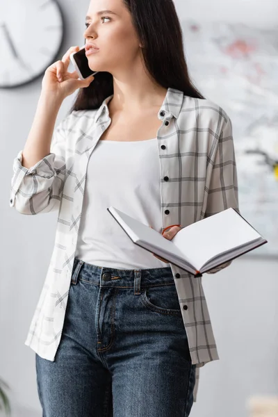 Young freelancer looking away while holding blank notebook and talking on mobile phone — Stock Photo