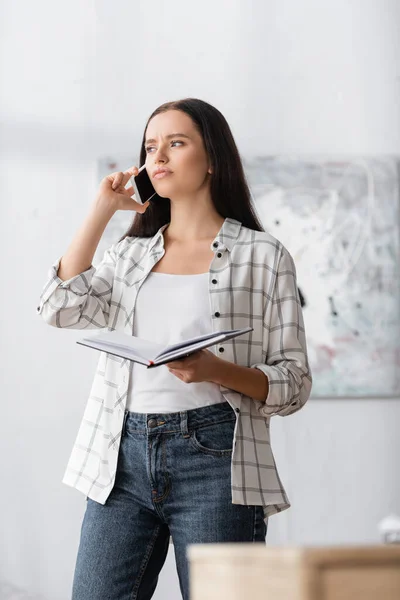 Nachdenklicher Freiberufler mit Notizbuch, der wegschaut, während er auf dem Handy im verschwommenen Vordergrund spricht — Stockfoto