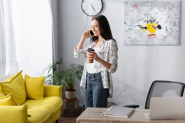 Smiling freelancer holding coffee to go while talking on mobile phone — Stock Photo