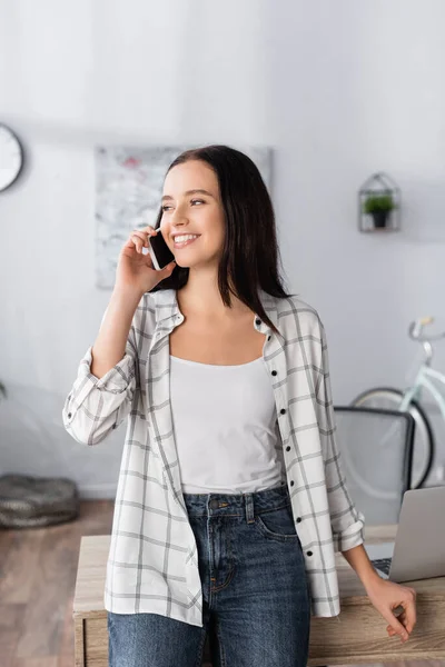 Mujer feliz hablando en el teléfono inteligente mientras mira hacia fuera en casa - foto de stock