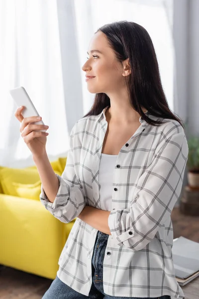 Joven mujer sonriendo mientras chatea en el teléfono inteligente y mirando hacia fuera en casa - foto de stock