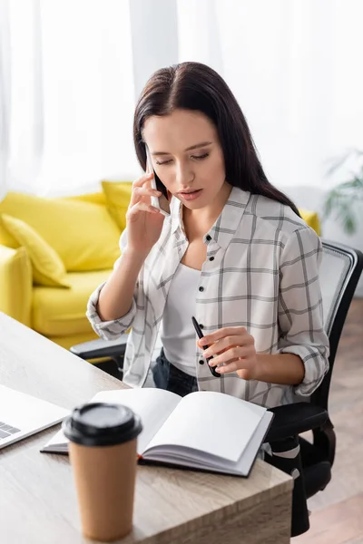 Freelancer talking on smartphone while holding pen near notebook and coffee to go on blurred foreground — Stock Photo