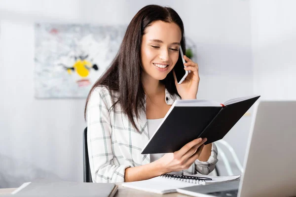 Cheerful freelancer talking on smartphone while holding notebook near laptop on blurred foreground — Stock Photo