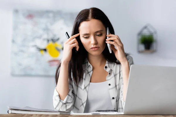 Aufgebrachte Freiberuflerin spricht auf Smartphone und hält Stift in der Nähe von Laptop auf verschwommenem Vordergrund — Stockfoto