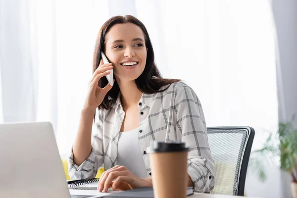 Alegre freelancer mirando hacia otro lado mientras habla en el teléfono inteligente cerca de la computadora portátil y el café para ir en primer plano borrosa - foto de stock