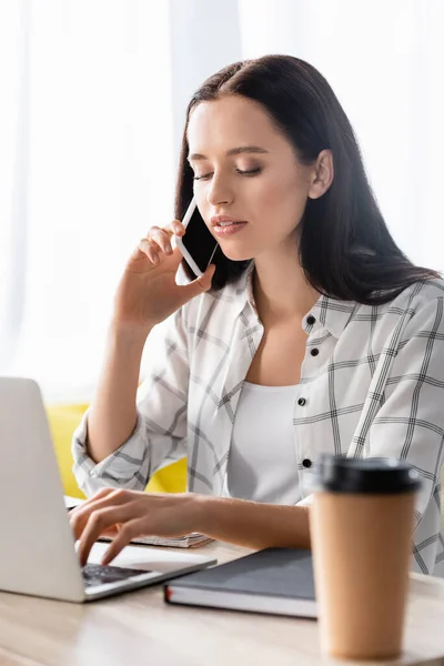 Young freelancer typing on laptop while talking on smartphone near coffee to go on blurred foreground — Stock Photo