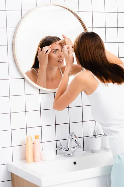Mujer joven insatisfecha tocando la cara mientras está de pie cerca del espejo en el baño - foto de stock