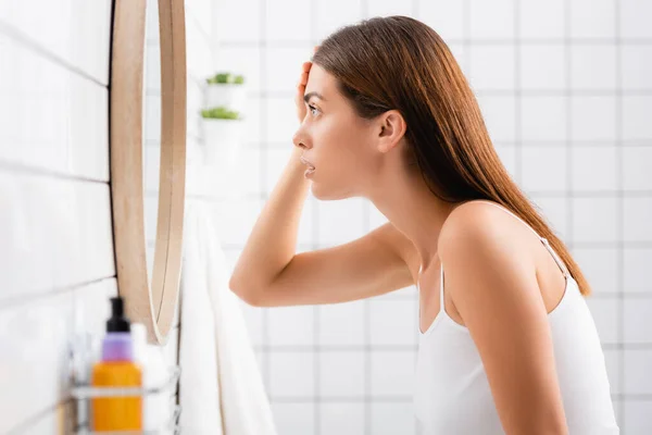 Seitenansicht einer besorgten jungen Frau, die beim Blick in den Spiegel die Stirn berührt — Stockfoto