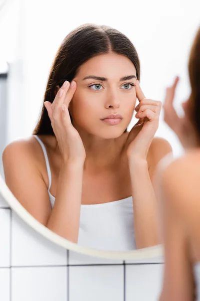 Junge Frau blickt in Spiegel und berührt Gesicht im Badezimmer, verschwommener Vordergrund — Stockfoto